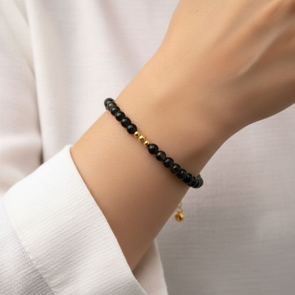 Vue rapprochée du poignet d'une personne portant le bracelet en onyx noire. Le bijou, composé de petites perles sombres et brillantes, entoure délicatement le poignet. Les trois perles dorées centrales sont bien visibles, apportant une touche de lumière contrastée sur la peau. La personne porte une chemise blanche légère, renforçant le style sobre et chic de l'accessoire.