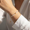 Gros plan du poignet d'une personne portant un bracelet fin en pierre de lune aux reflets blancs et nacrés. Les petites perles rondes captent la lumière naturelle, créant un contraste doux avec les trois perles dorées centrales. Le bijou est porté avec un vêtement blanc immaculé, soulignant l'élégance minimaliste et la clarté de la pierre.
