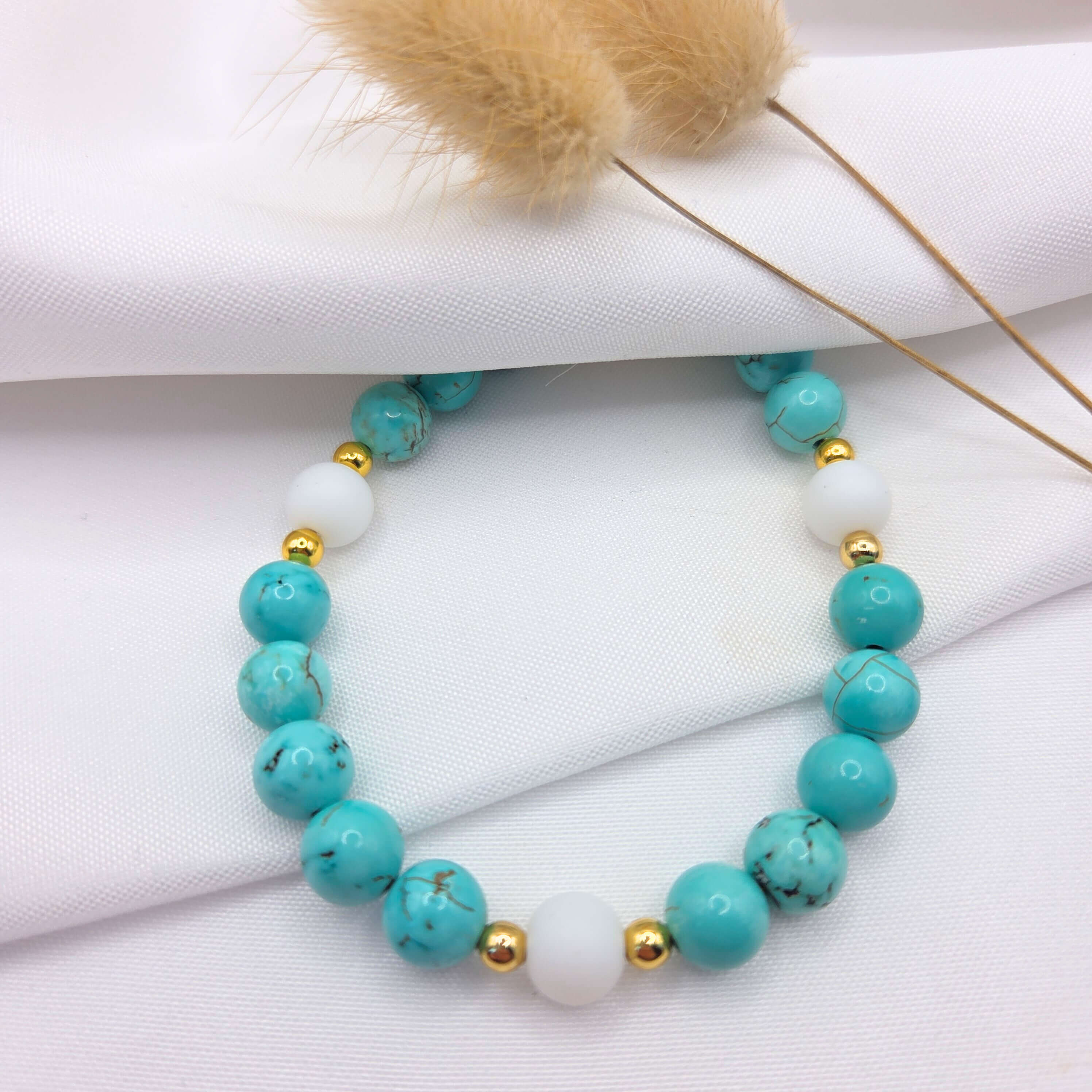 Bracelet en perles de turquoise et perles blanches.

Le bracelet est présenté à plat sur un tissu blanc texturé. Il est composé de perles rondes en turquoise véritable (ou imitation turquoise) montrant des nervures brunes naturelles, alternées avec trois perles blanches mates plus larges. Chaque perle blanche est entourée de petites perles d'espacement dorées brillantes. En arrière-plan, deux tiges de fleurs séchées  apportent une touche décorative douce et nature