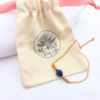 Bracelet délicat avec une pierre centrale en Lapis Lazuli bleu profond, entourée de perles dorées sur une fine chaîne plaqué or. Le bijou repose sur un petit sac de rangement en coton beige avec le logo noir M-ily Glow Up.
