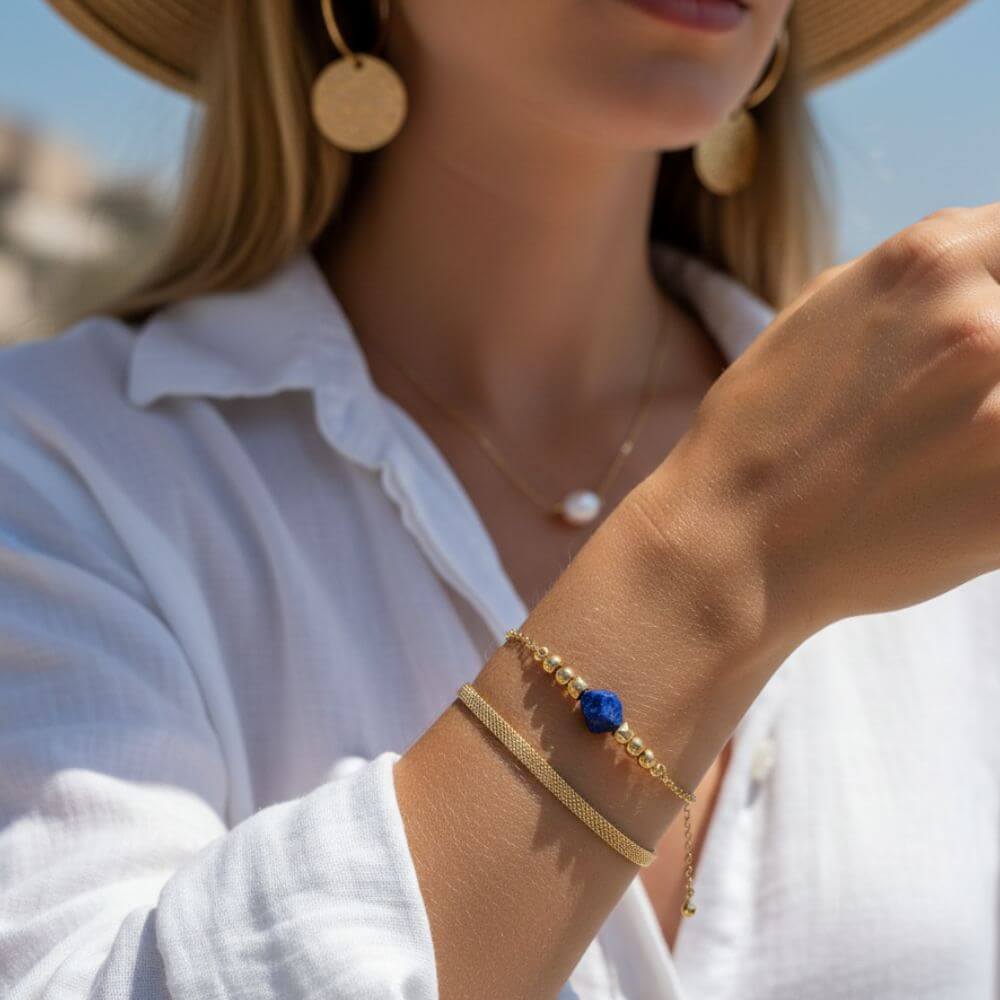 Gros plan sur une femme portant un duo de bracelets dorés, l'un en maille fine et l'autre orné d'une pierre de Lapis Lazuli bleue brute.
