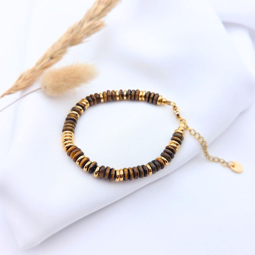 Bracelet Iris composé de perles rondelles en pierre naturelle Œil de Tigre marron et d'intercalaires dorés, posé sur un tissu blanc avec une touche végétale.