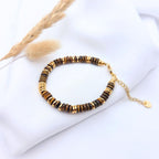 Bracelet Iris composé de perles rondelles en pierre naturelle Œil de Tigre marron et d'intercalaires dorés, posé sur un tissu blanc avec une touche végétale.