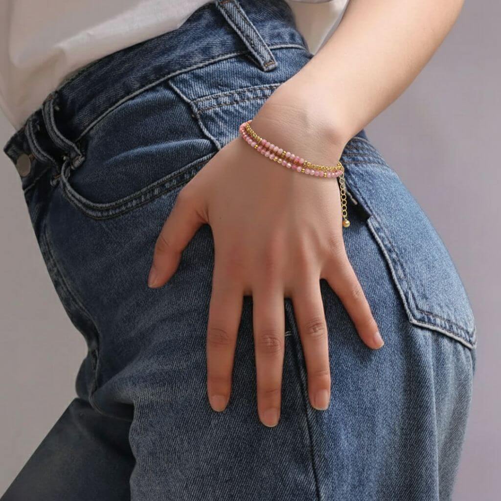 Gros plan sur le poignet d'une personne portant un bracelet délicat à deux rangs, composé de petites perles roses de rhodonite et de billes dorées. La main repose sur la hanche, vêtue d'un jean bleu délavé et d'un haut blanc.
