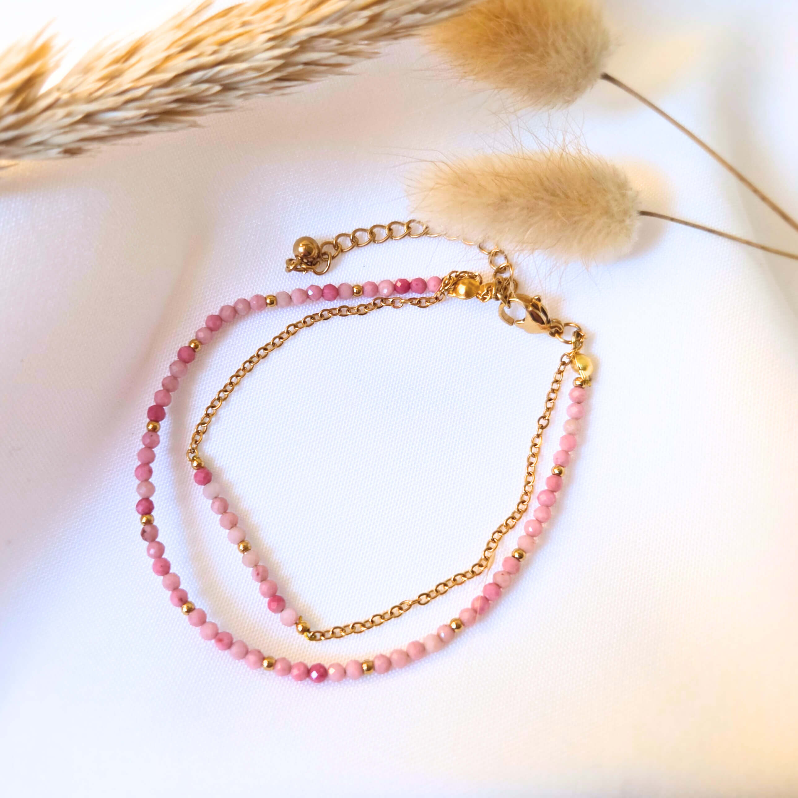 Bracelet double rang délicat composé d'une fine chaîne dorée et d'un fil de petites perles de rhodonite dans des nuances de rose poudré et de vieux rose. Le bijou est muni d'un fermoir mousqueton et d'une chaînette de réglage dorés, posés sur un tissu blanc avec des graminées séchées en arrière-plan.