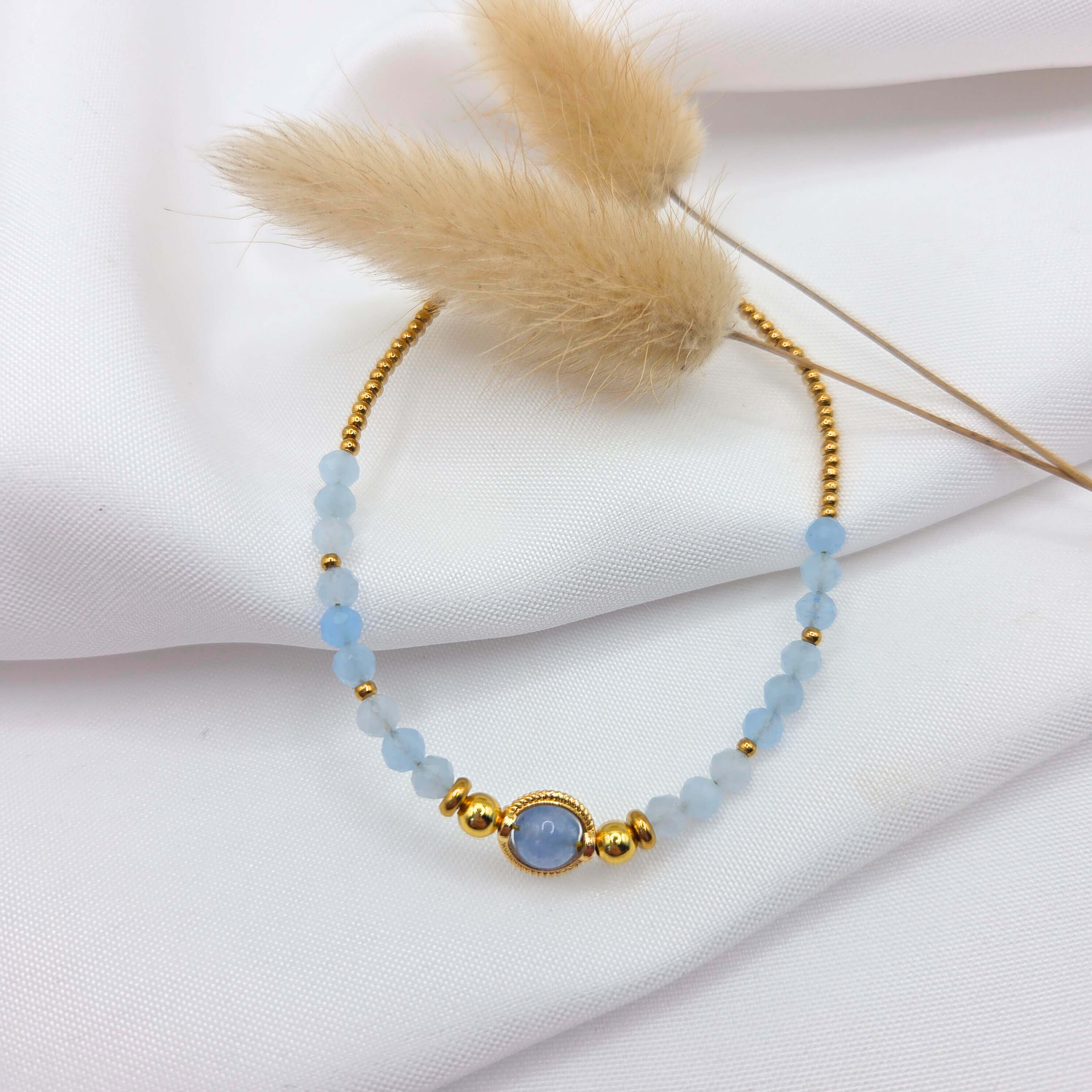 Un bracelet fin disposé sur un tissu blanc texturé, décoré de deux plumeaux de graminées séchées. Le bijou se compose de petites perles dorées sur la partie supérieure et de perles facettées d'aigue marine bleu clair translucides sur la partie inférieure. Au centre, une perle ronde d'aigue marine plus large est sertie dans un médaillon doré travaillé.