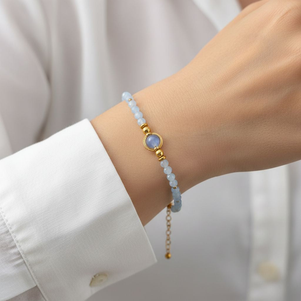 Gros plan du même bracelet porté au poignet d'une personne vêtue d'une chemise blanche. La pierre centrale d'aigue marine bleue et les perles facettées bleu clair ressortent sur la peau. On aperçoit la chaînette de réglage dorée qui pend légèrement sous le poignet. La main est partiellement visible, posée avec élégance.