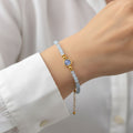 Gros plan du même bracelet porté au poignet d'une personne vêtue d'une chemise blanche. La pierre centrale d'aigue marine bleue et les perles facettées bleu clair ressortent sur la peau. On aperçoit la chaînette de réglage dorée qui pend légèrement sous le poignet. La main est partiellement visible, posée avec élégance.