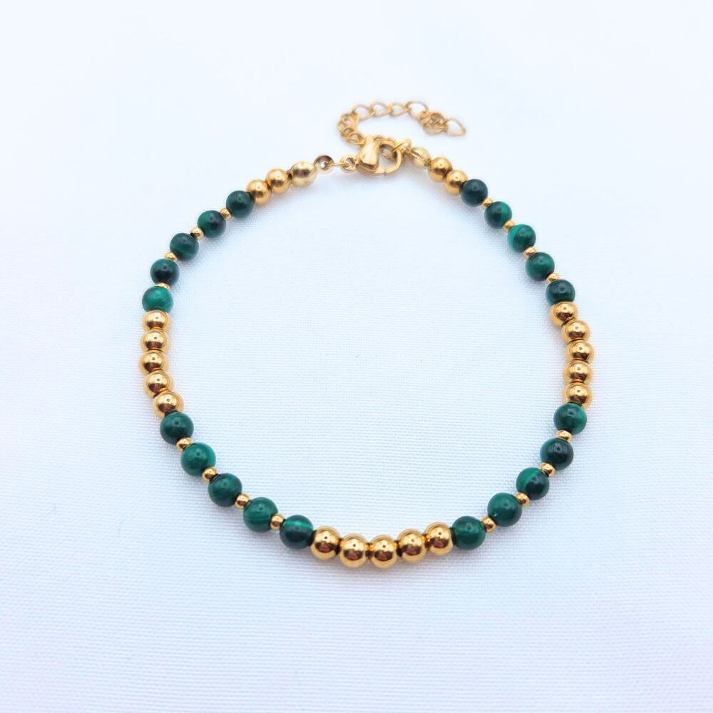 Bracelet fin composé de perles de malachite verte veinée et de billes dorées en acier inoxydable, posé sur un fond blanc texturé.