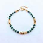 Bracelet fin composé de perles de malachite verte veinée et de billes dorées en acier inoxydable, posé sur un fond blanc texturé.