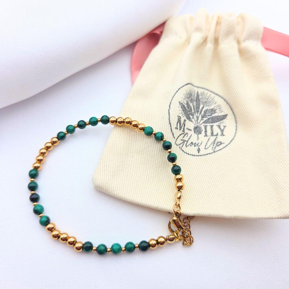 Bracelet fin en perles de malachite verte et perles dorées, posé sur un pochon en lin blanc de la marque M-ily Glow Up.