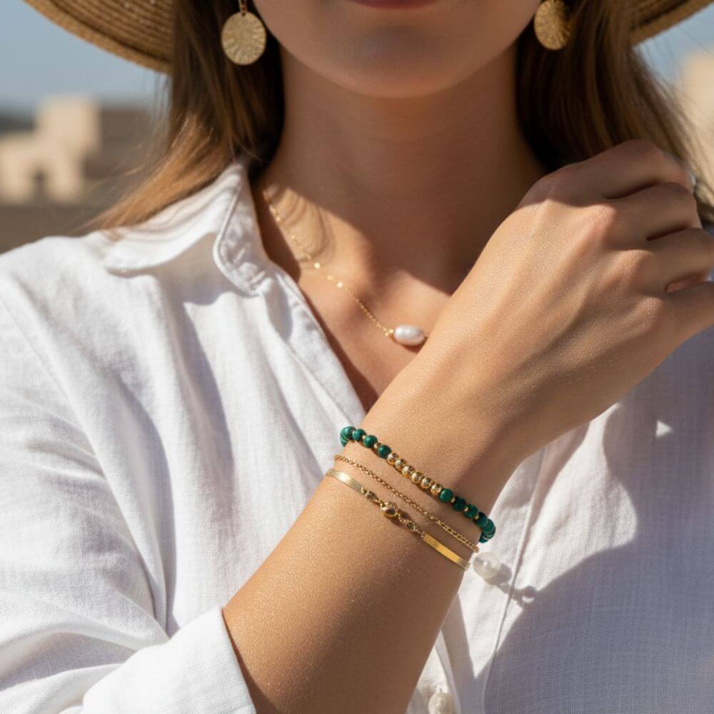 Femme portant une chemise en lin blanc et des bijoux dorés : collier avec perle nacrée et accumulation de bracelets en or et perles de malachite verte.