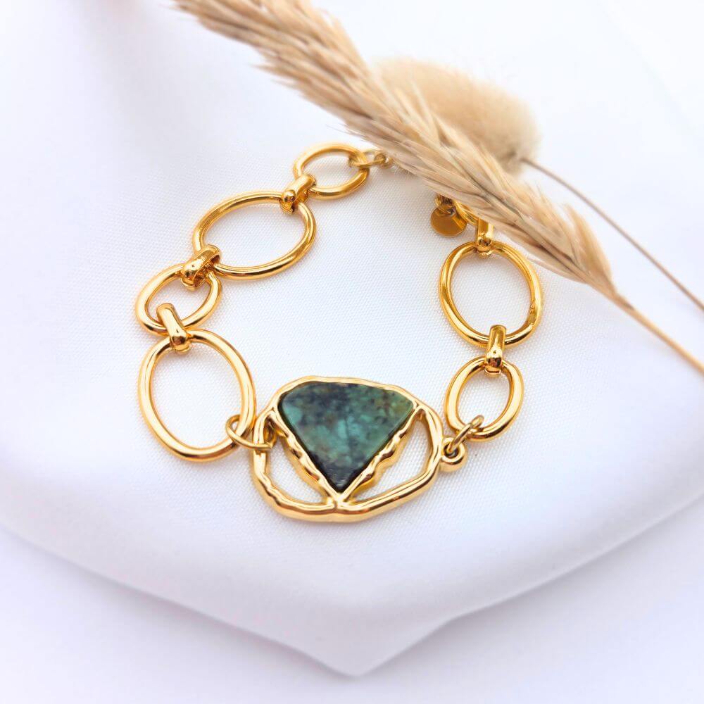 Bracelet Sélène composé d'une pierre turquoise africaine triangulaire au centre d'une chaîne à gros maillons dorés, posé sur un tissu blanc avec un épi de blé.