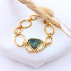 Bracelet Sélène composé d'une pierre turquoise africaine triangulaire au centre d'une chaîne à gros maillons dorés, posé sur un tissu blanc avec un épi de blé.