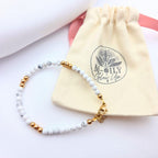 Bracelet fin en perles de howlite blanche marbrée et perles dorées, posé devant son pochon en toile beige marqué "M-ILY Glow Up".