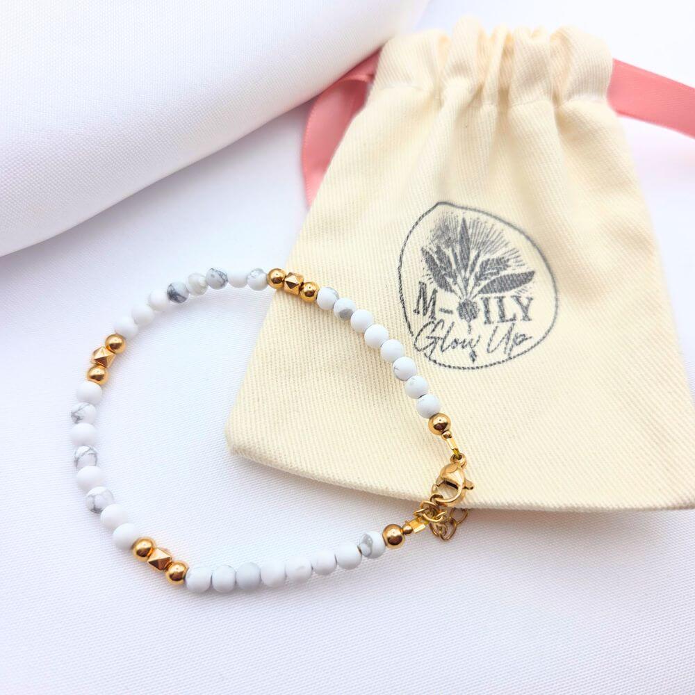 Bracelet fin en perles de howlite blanche marbrée et perles dorées, posé devant son pochon en toile beige marqué "M-ILY Glow Up".
