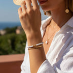 Femme portant un ensemble de bijoux dorés et un bracelet en perles de howlite blanche, vêtue d'une chemise en lin blanc en extérieur ensoleillé.