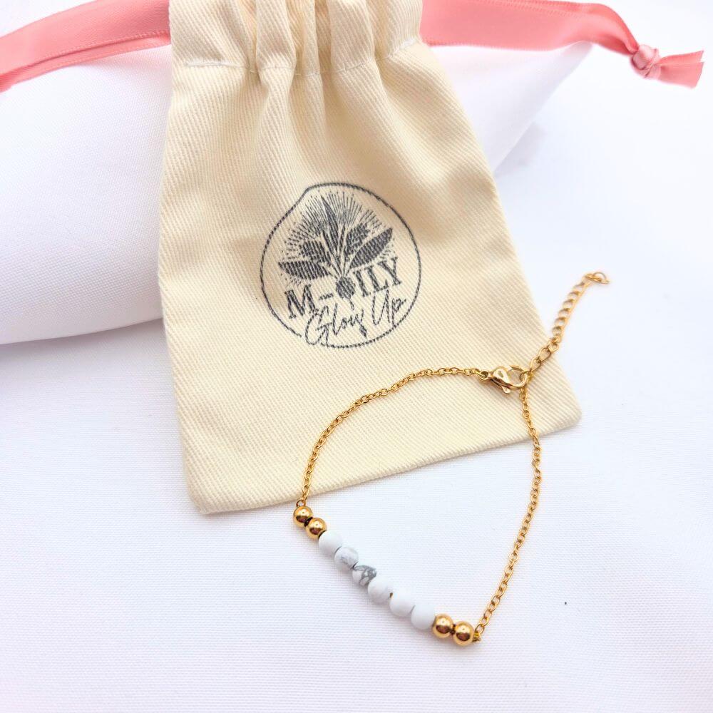 Bracelet fin en chaîne dorée avec perles de howlite blanche et marbrée, posé devant son pochon en coton beige logoté M-ILY Glow Up.