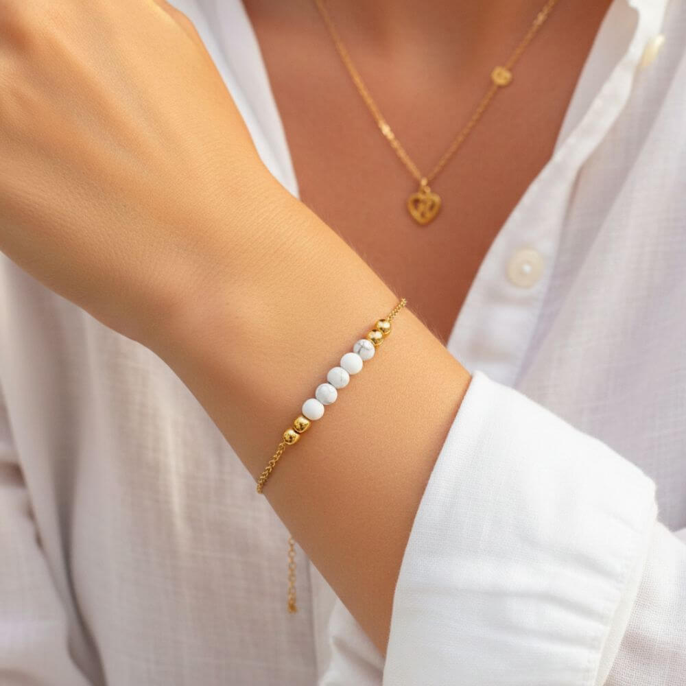 Bracelet fin doré pour femme orné de perles en Howlite blanche et billes dorées, porté au poignet avec une chemise blanche.