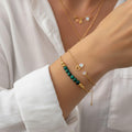 Femme portant une superposition de bracelets dorés, l'un avec des perles de malachite verte et l'autre avec un pendentif cœur et des pierres d'opale.