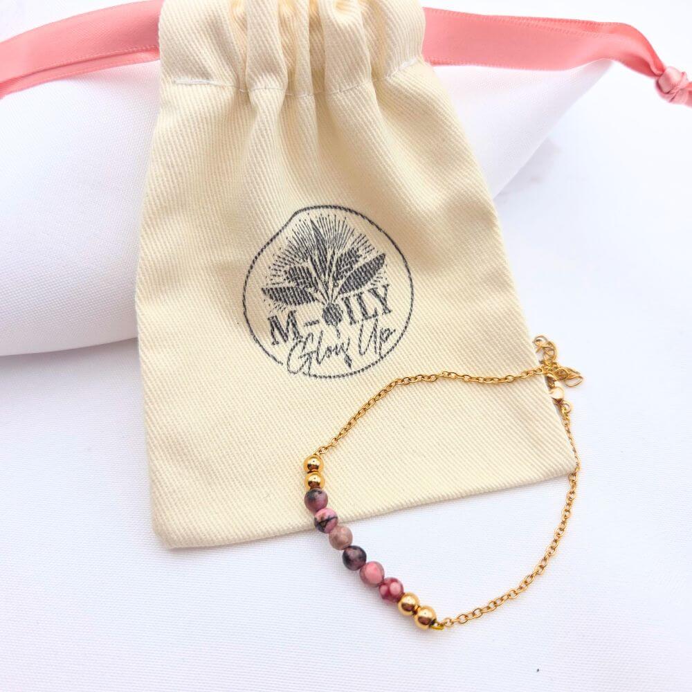 Bracelet fin en chaîne dorée orné de perles de rhodonite roses et noires, posé devant son pochon en coton naturel logoté "M-ILY Glow Up".