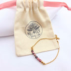 Bracelet fin en chaîne dorée orné de perles de rhodonite roses et noires, posé devant son pochon en coton naturel logoté "M-ILY Glow Up".