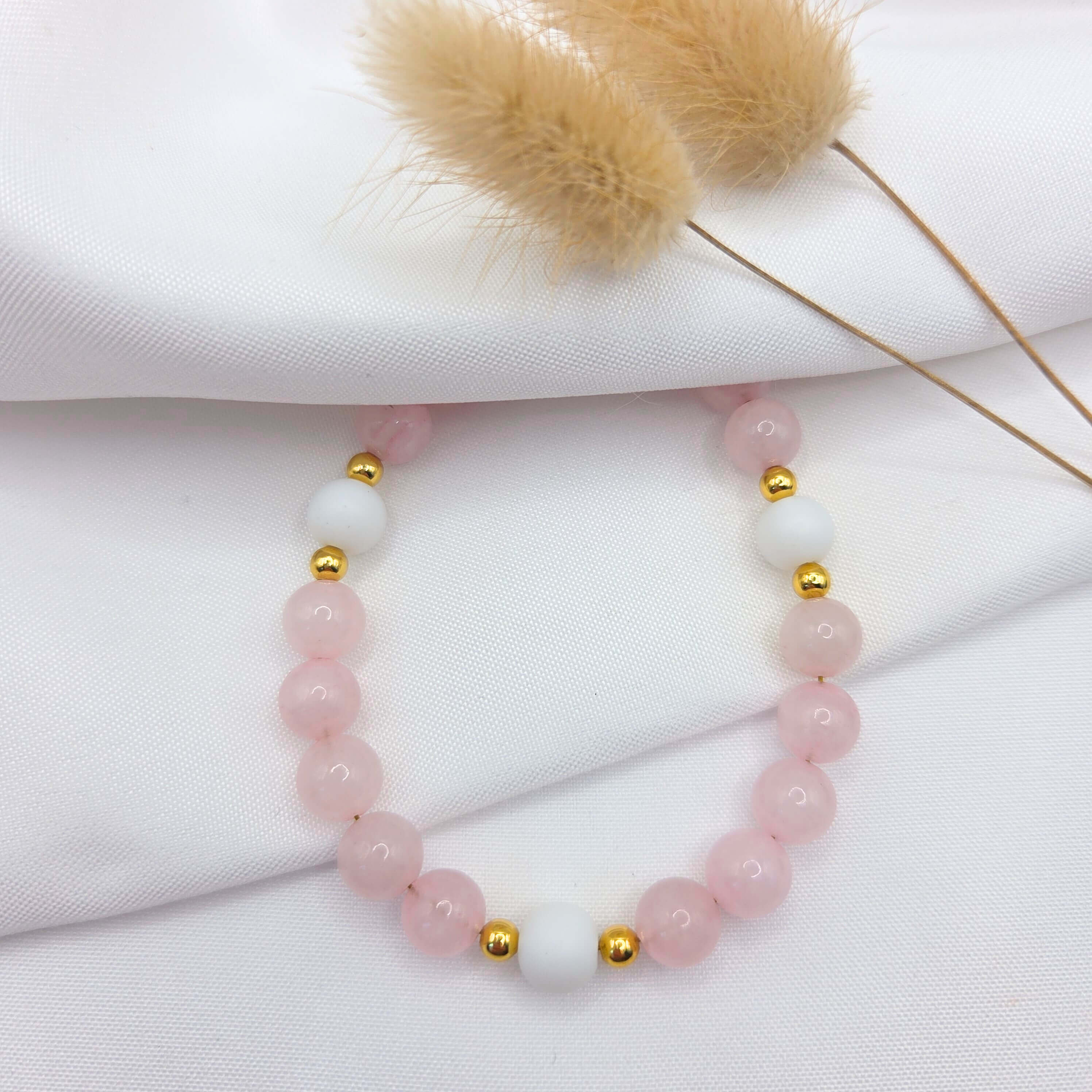 Vue de dessus d'un bracelet en perles de quartz rose poli, disposé sur un tissu blanc drapé. Le bijou intègre trois perles d'un blanc mat, chacune encadrée par de petites billes dorées brillantes. Deux plumeaux de graminées séchées décorent délicatement le haut de la scène.