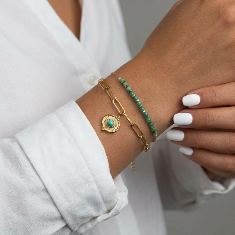 Une photo close-up d'un poignet féminin portant le bracelet Alma. Le bracelet double rang se compose d'une fine chaîne en acier inoxydable doré ornée de perles de jade rondes et polies, et d'une chaîne plus épaisse avec un pendentif rond et gravé serti d'une pierre de turquoise centrale. La personne porte une chemise en lin blanche et a une manucure soignée avec du vernis à ongles blanc. L'arrière-plan est légèrement flou.