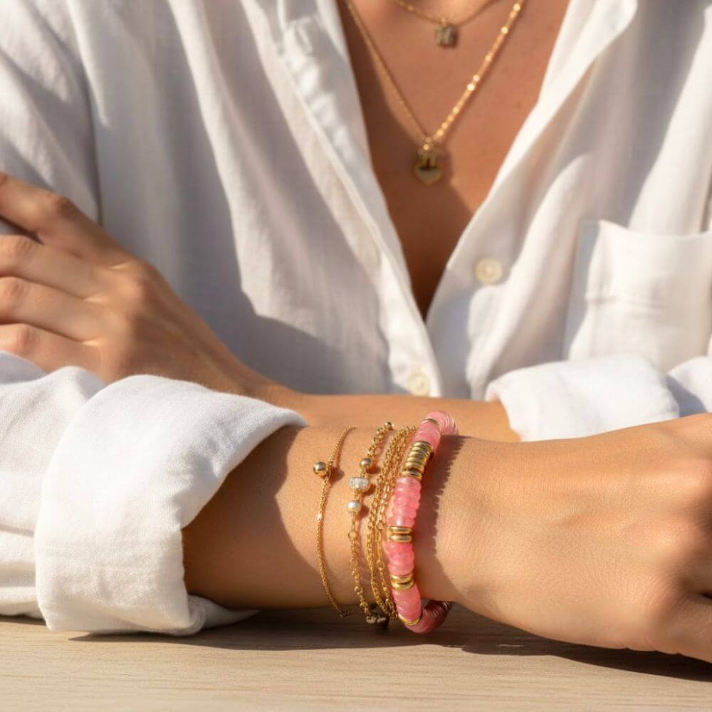 Bracelet Iris avec perles en quartz rose véritable porté en accumulation avec des joncs dorés sur un poignet ensoleillé.
