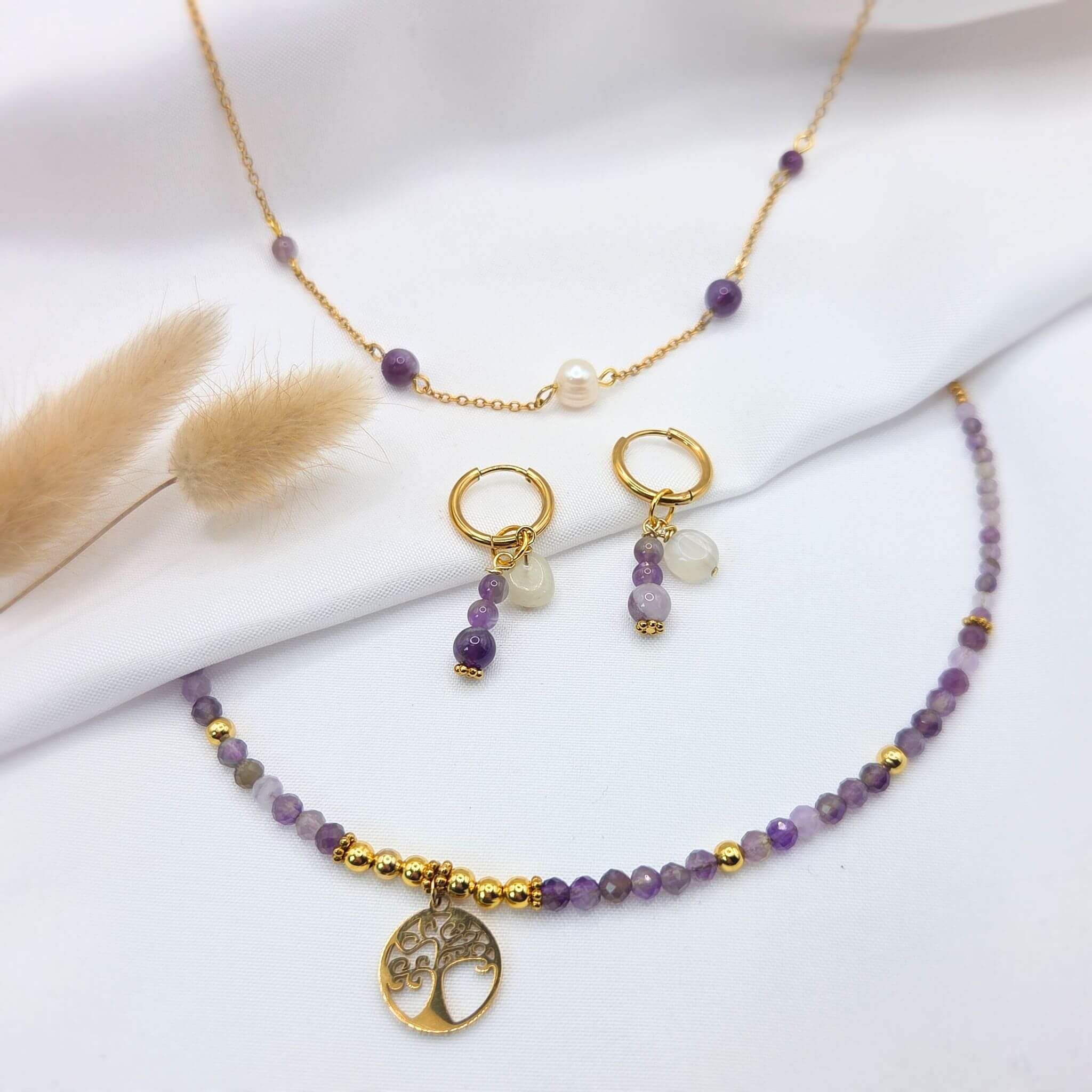 Composition à plat d'une parure de bijoux dorés et améthyste sur un tissu blanc drapé. L'ensemble comprend un collier de perles d'améthyste facettées avec un pendentif Arbre de Vie, une fine chaîne dorée ponctuée de perles violettes et d'une perle de culture blanche, et une paire de boucles d'oreilles créoles ornées de pampilles en pierres naturelles.