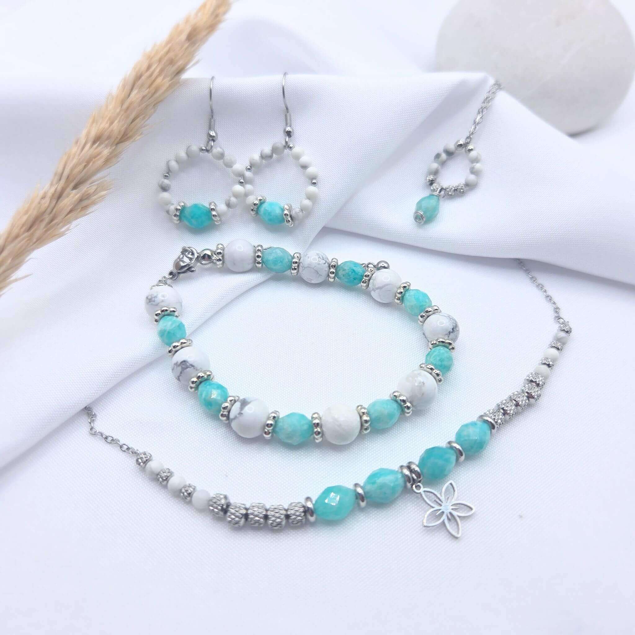 Parure de bijoux artisanaux de la collection GIA par M-ily Glow Up. L'ensemble comprend un bracelet en perles, un collier avec pendentif fleur argenté, des boucles d'oreilles créoles et un sautoir assortie, tous composés de Howlite blanche et d'Amazonite turquoise.