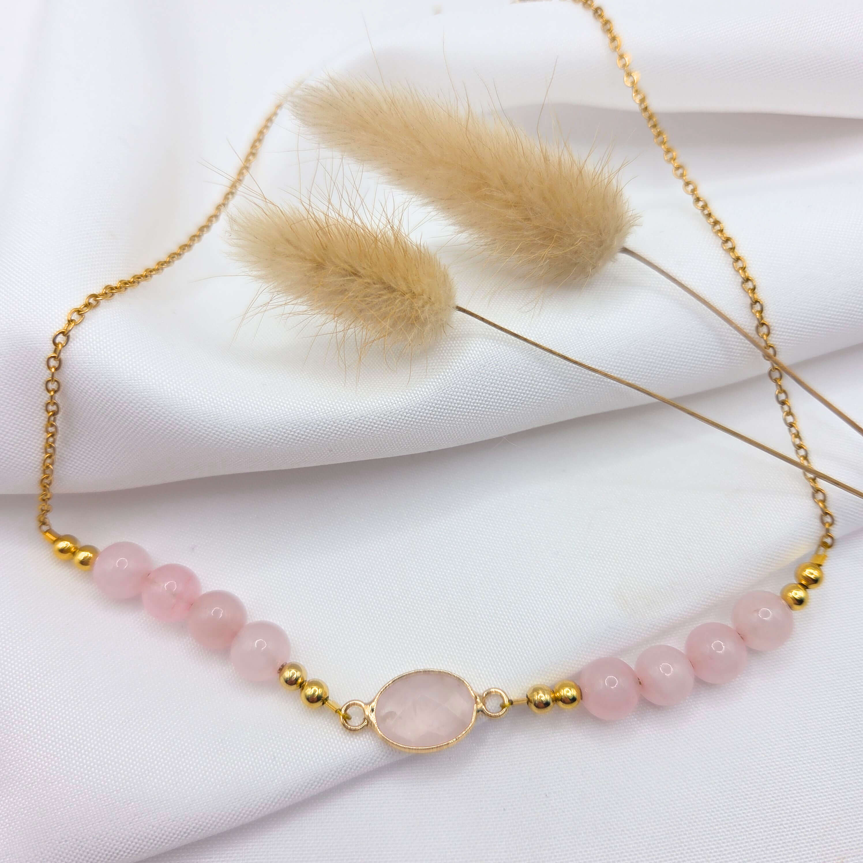Un collier délicat en acier inoxydable doré posé sur un tissu blanc fluide. Le bijou est orné de perles rondes en quartz rose pâle de chaque côté d'un pendentif central ovale en pierre rose sertie d'or. Deux tiges de fleurs séchées de couleur beige naturel sont disposées en arrière-plan, renforçant l'aspect doux et bohème de la composition.