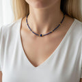 Gros plan sur le cou d'une personne portant un collier de perles bleues fines et élégantes. Le collier se compose d'une multitude de petites perles de lapis-lazuli bleu nuit, parsemées de quelques perles dorées brillantes. Au centre, une perle bleue légèrement plus grosse est encadrée par deux perles dorées. La personne porte un vêtement blanc épuré qui fait ressortir les couleurs contrastées du bijou.