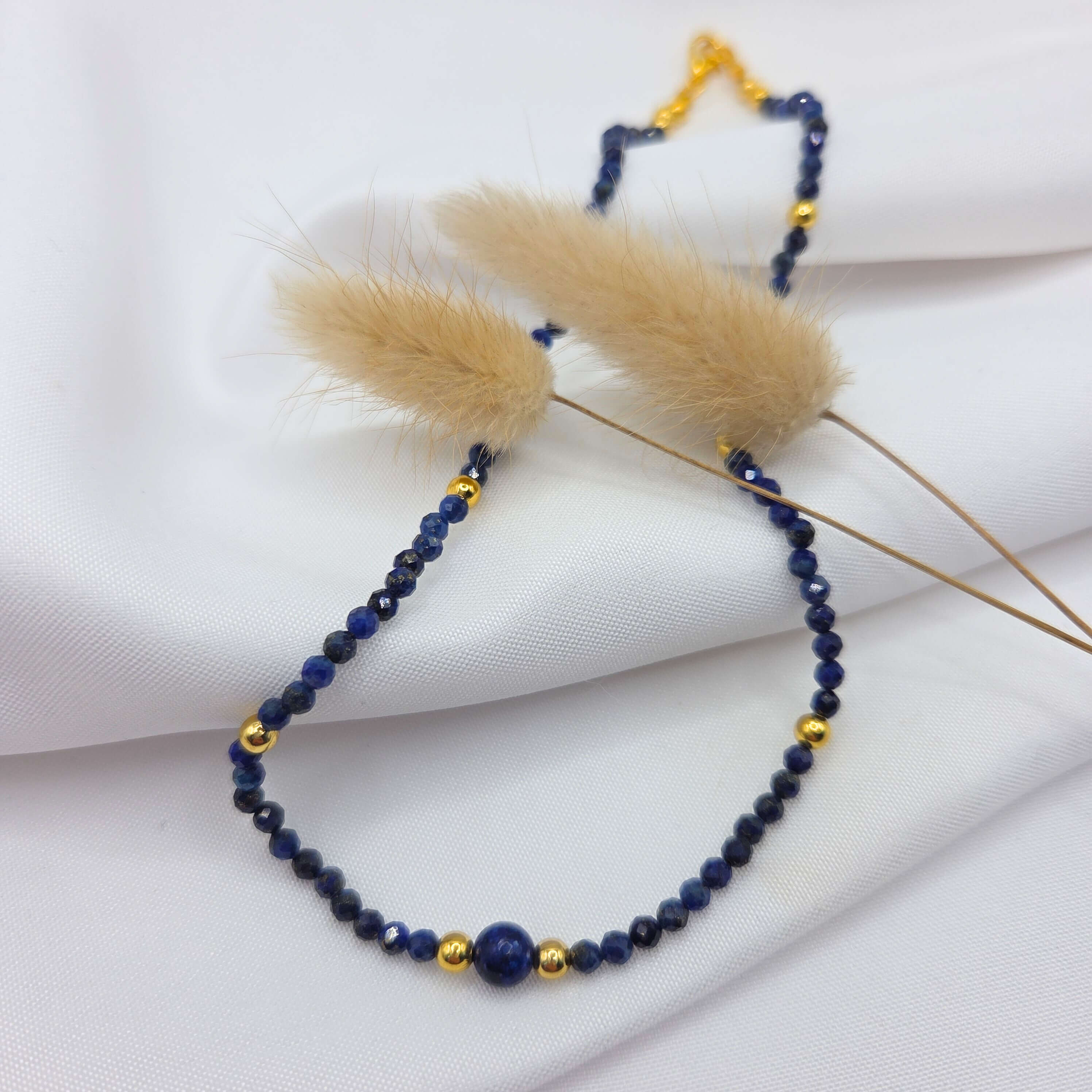 Un collier délicat composé de petites perles de lapis-lazuli d'un bleu profond, ponctuées de perles dorées lisses et brillantes. Le bijou est élégamment disposé sur un tissu blanc fluide et drapé. Deux brins d'herbe de la pampa séchés, aux plumeaux beiges et duveteux, traversent la composition, apportant une touche douce et naturelle à l'ensemble.