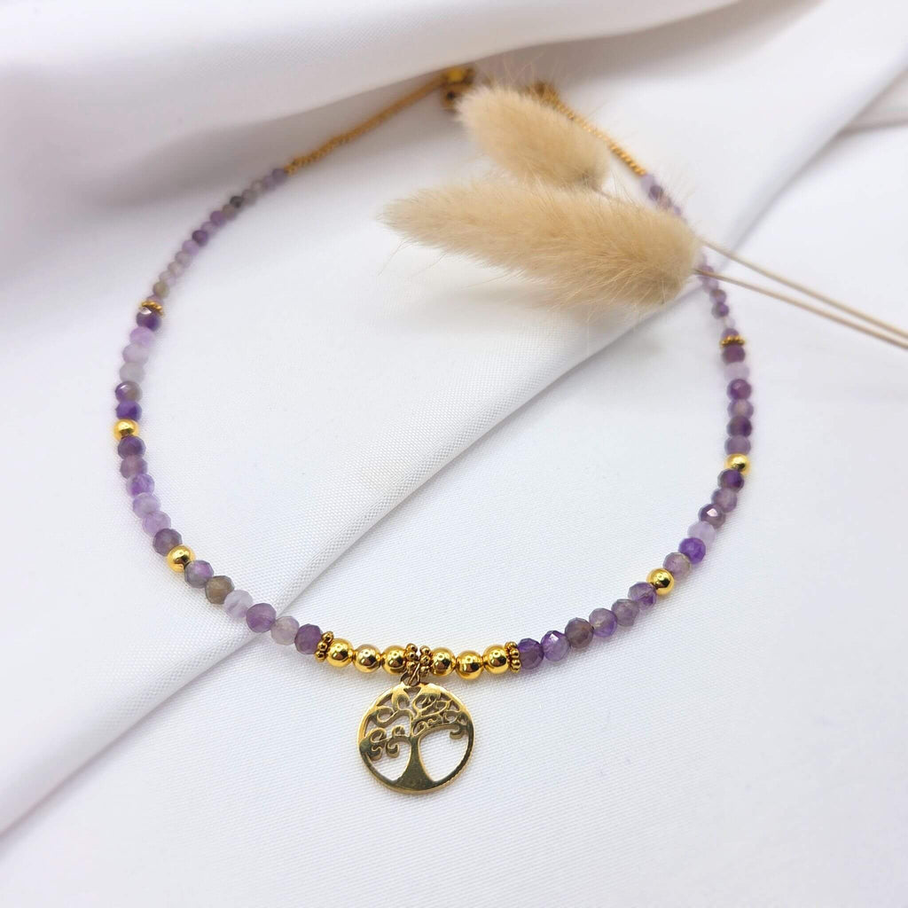 Collier artisanal composé de petites perles d'améthyste violettes facettées et de perles dorées, orné en son centre d'un pendentif rond représentant un "Arbre de Vie" en métal doré, posé sur un tissu blanc drapé.