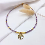 Collier artisanal composé de petites perles d'améthyste violettes facettées et de perles dorées, orné en son centre d'un pendentif rond représentant un "Arbre de Vie" en métal doré, posé sur un tissu blanc drapé.