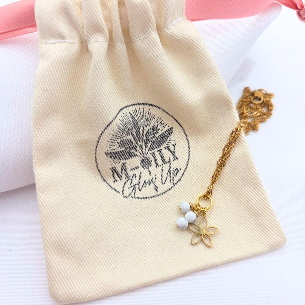 Collier doré à mailles torsadées avec pendentif fleur évidée et perles blanches, posé sur un pochon en coton beige marqué du logo "M-ily Glow Up".