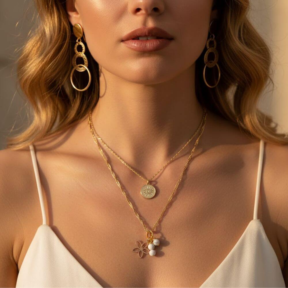 Superposition de colliers dorés : une chaîne fine avec médaille soleil gravée et un collier torsadé avec charms fleur ajourée et perles marbrées blanches sur un débardeur blanc