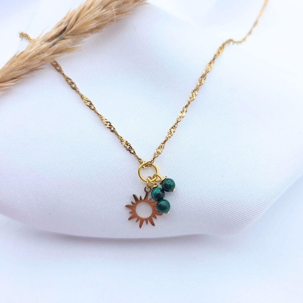 Collier en chaîne torsadée dorée avec pendentif soleil évidé et trois perles de malachite verte sur fond blanc.