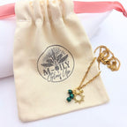 Collier doré avec pendentif soleil et perles vertes de malachite, posé sur son pochon en coton naturel logotypé "M-ILY Glow Up".