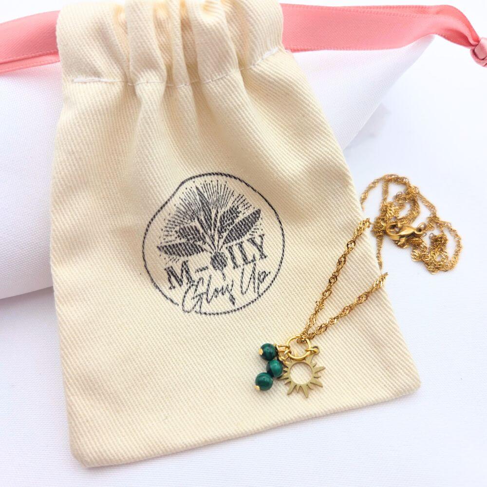 Collier doré avec pendentif soleil et perles vertes de malachite, posé sur son pochon en coton naturel logotypé "M-ILY Glow Up".