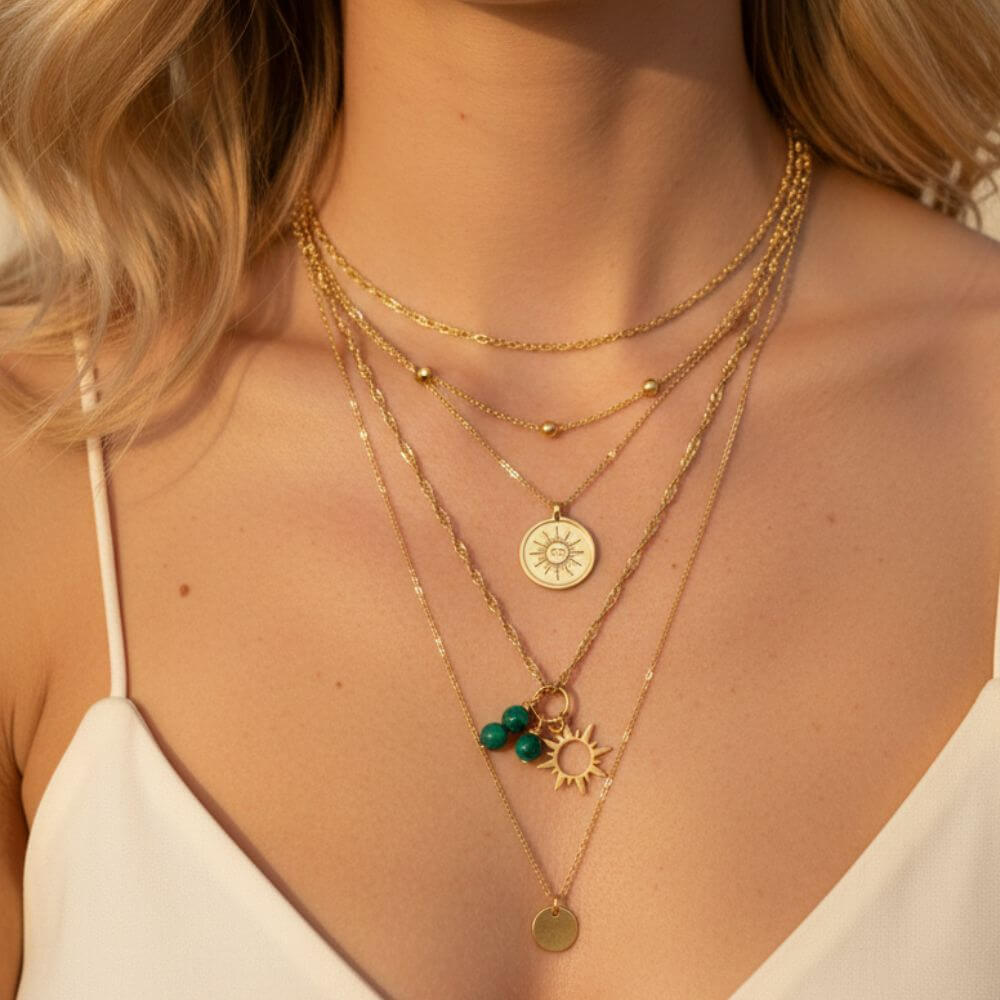 Gros plan sur une femme portant une superposition de cinq colliers dorés, comprenant une chaîne à billes, un médaillon soleil gravé, des perles vertes en malachite et un pendentif disque minimaliste.