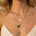 Gros plan sur une femme portant une superposition de cinq colliers dorés, comprenant une chaîne à billes, un médaillon soleil gravé, des perles vertes en malachite et un pendentif disque minimaliste.