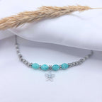 Photo en gros plan d'un collier artisanal "Gia" présenté sur un tissu blanc fluide. Le bijou se compose de perles d'Amazonite facettées de couleur turquoise au centre, entourant un pendentif en forme de fleur argentée délicate. Le reste du collier alterne des perles en argent ciselé et de petites perles de Howlite blanche. Un épi de blé séché est posé en arrière-plan pour une ambiance bohème et épurée.