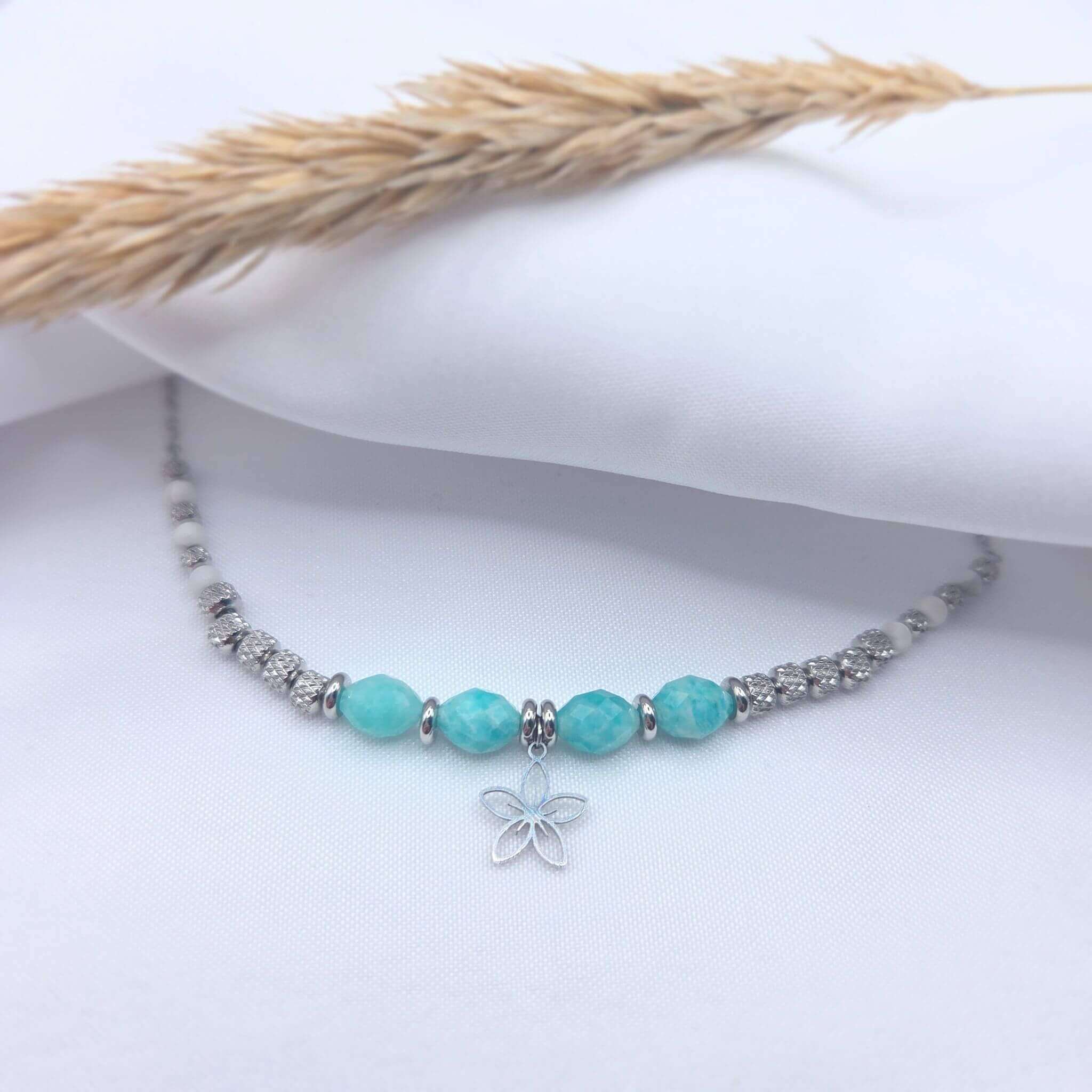 Photo en gros plan d'un collier artisanal "Gia" présenté sur un tissu blanc fluide. Le bijou se compose de perles d'Amazonite facettées de couleur turquoise au centre, entourant un pendentif en forme de fleur argentée délicate. Le reste du collier alterne des perles en argent ciselé et de petites perles de Howlite blanche. Un épi de blé séché est posé en arrière-plan pour une ambiance bohème et épurée.