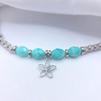 Gros plan sur un collier en argent présentant quatre perles d'Amazonite turquoise facettées, séparées par de petits anneaux argentés, avec un pendentif central en forme de fleur délicate posé sur un tissu blanc.