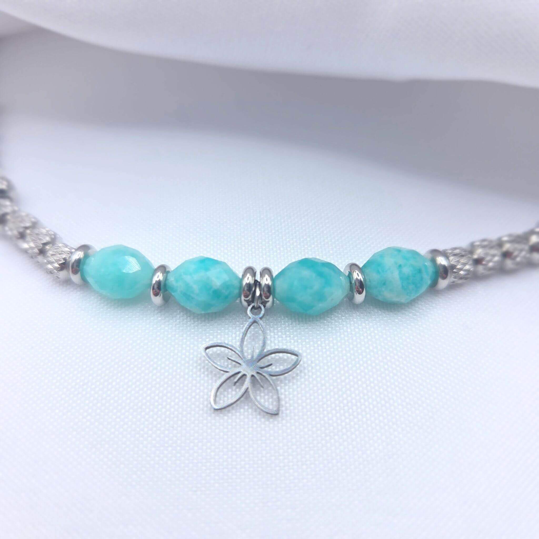 Gros plan sur un collier en argent présentant quatre perles d'Amazonite turquoise facettées, séparées par de petits anneaux argentés, avec un pendentif central en forme de fleur délicate posé sur un tissu blanc.