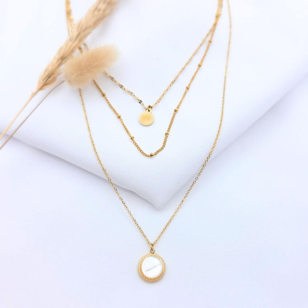 Collier Rivage avec pendentif rectangulaire aux bords irréguliers et martelés, chaîne dorée fine posée sur un tissu blanc avec un épi de blé.