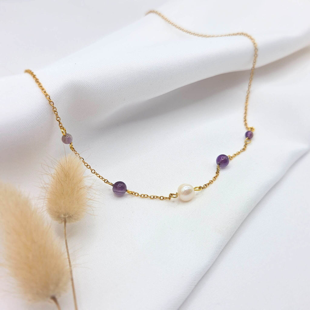 Collier fin en chaîne dorée délicate, orné d'une perle d'eau douce centrale entourée de quatre perles d'améthyste violettes disposées de manière symétrique. Le bijou est mis en valeur sur un tissu de soie blanc drapé, accompagné de quelques tiges de fleurs séchées duveteuses au premier plan.