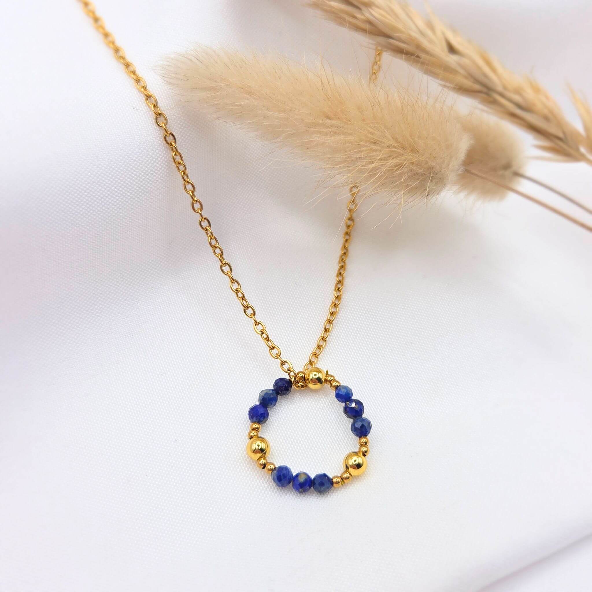 Collier à chaîne fine dorée orné d'un pendentif en forme d'anneau composé de perles de lapis-lazuli bleu profond facettées, alternées avec des billes d'or lisses. Le bijou est mis en scène sur un tissu blanc immaculé, accompagné de graminées séchées pour une ambiance naturelle et raffinée.